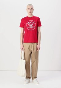 T-shirt en coton rouge avec un design de logo circulaire, pantalon beige, baskets blanches et sac fourre-tout beige. Design simple et coupe décontractée.