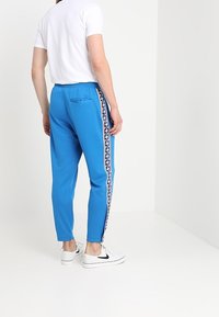 Muž stojící v bílých teniskách, bílém tričku s krátkým rukávem a jasně modrých teplácích s proužky na bocích a kapsou na zádech.