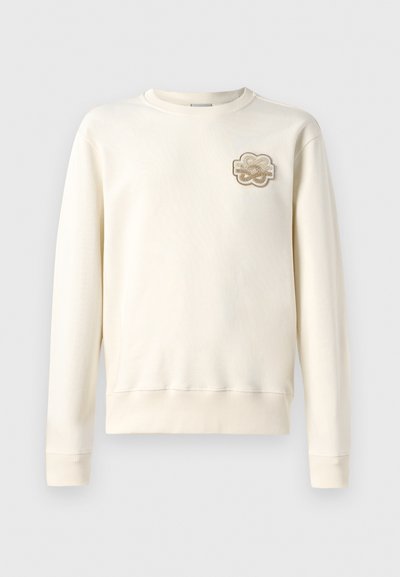 Gaiši bēšs džemperis ar apaļu kakla izgriezumu, garām piedurknēm un ribotām aprocēm. Uz kreisā krūtīm ir dekoratīvs izšūts plāksteris.