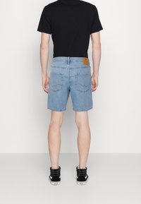 Ljusblå denimshorts med rak skärning, utrustade med en brun läderlapp på baksidan av midjan och två bakfickor. Svarta sneakers synliga.