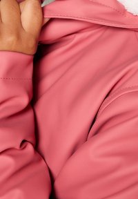 Rosa ytterkläder med en slät, glansig yta, med en mjuk, fleece-fodrad krage och sydda detaljer. Hand som greppar tyget.