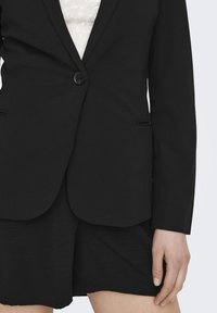 Osoba má na sobě černou blazeru přes krajkový top, doplněnou černými šortkami. Pozadí je neutrální, což zdůrazňuje detaily outfitu.