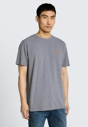 Mężczyzna ubrany w prostą szarą koszulkę z krótkim rękawem z małym czerwonym napisem na piersi, stojący na jasnym tle.