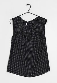 Top nero senza maniche con pois bianchi, scollatura rotonda e un piccolo dettaglio a forma di chiave sul retro, appeso a un gancio nero.