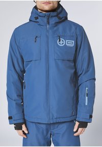 Blaue Außenjacke mit Kapuze, ausgestattet mit zwei Brusttaschen, seitlicher Reißverschlusstasche und einem Logo. Hergestellt aus strapazierfähigem, strukturiertem Material.