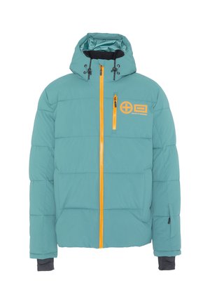 Mann trägt eine türkisfarbene Steppjacke mit gelbem Reißverschluss, farbblockierte Cargohose und Winterstiefel, steht und schaut seitlich.