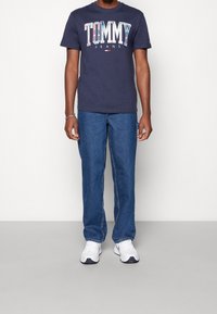 Marinblå bomulls-t-shirt med "TOMMY JEANS" tryckt i flerfärgade bokstäver, i kombination med blå denimjeans och vita sportskor.