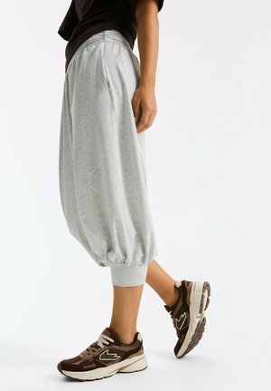 Person wearing loose gray cropped pants and brown sneakers standing on white background.