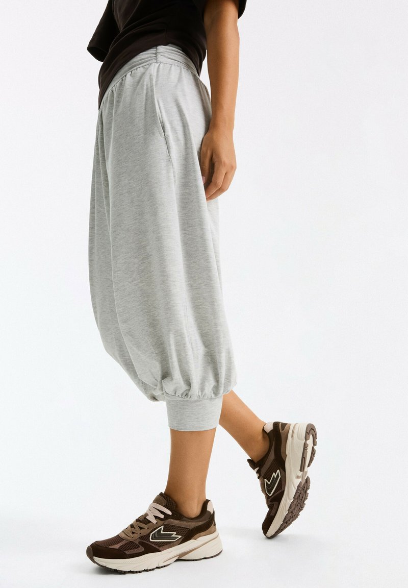 Personne portant un pantalon gris ample court et des baskets marron, debout sur fond blanc.