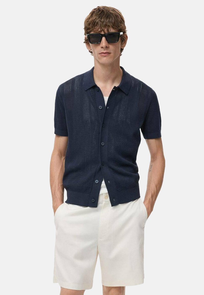 Cárdigan de punto de manga corta en color marino con cierre de botones, patrón texturizado y cuello clásico, combinado con pantalones cortos de color claro.