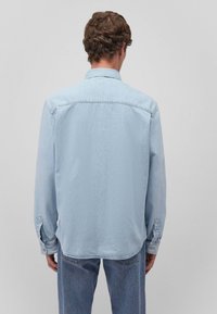 Camicetta in denim azzurro chiaro con maniche lunghe, polsini con bottoni, colletto classico e orlo curvo, che mostra una texture liscia e un design semplice.