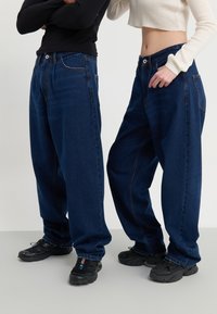 Två personer klädda i lösa mörkblå jeans och svarta sneakers, stående mot en enkel ljus bakgrund.