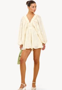 Monos de color crema con un patrón floral texturizado, escote en V profundo, ribete con volantes, mangas abullonadas, combinado con un bolso verde y calzado claro.