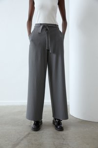 Pantalons larges gris avec des rayures verticales blanches, présentant une taille ceinturée et des poches, associés à des chaussures à lacets noires sur un sol en béton.