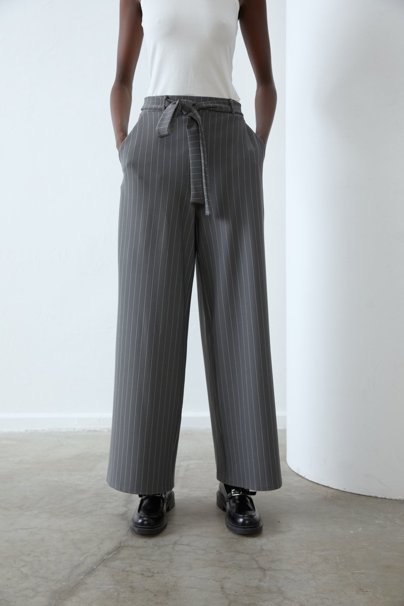 Pantalons larges gris avec des rayures verticales blanches, présentant une taille ceinturée et des poches, associés à des chaussures à lacets noires sur un sol en béton.
