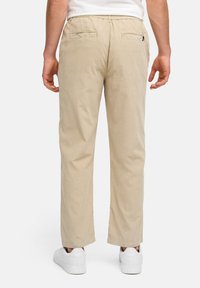 Pantalones beige con un corte relajado, cinturilla elástica y dos bolsillos traseros. Fabricados con una tela suave de textura lisa. Zapatillas blancas puestas.