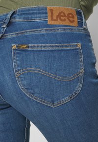 Blå denimjeans med en brun läderlapp märkt "Lee" på midjan, med gula sömmar och en rundad bakfickdesign.