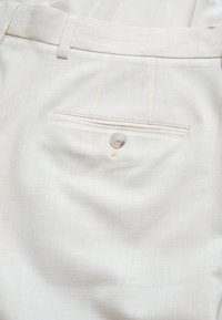 Beige Hose aus leichtem Stoff mit glatter Textur, klassischen Taschen und einem einzelnen Knopfverschluss an der hinteren Tasche.