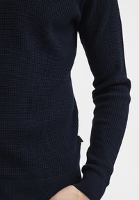 Pulover tricotat de culoare albastru închis, prezentând un model texturat în formă de vafe, cu manșete și tiv ribbate. Materialul pare cald și elastic, potrivit pentru purtarea de zi cu zi.