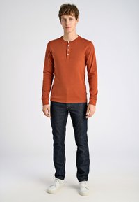 Pitkähihainen, ruosteenvärinen henley-paita, jossa on kolme nappia, yhdistettynä tummansinisiin slim fit -farkkuihin ja valkoisiin tennareihin. Yksinkertainen muotoilu ja tekstuuri.