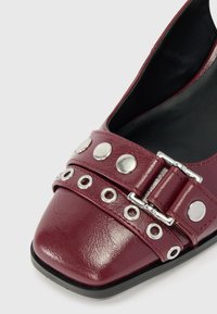 Scarpa in pelle bordeaux con punta squadrata, dotata di una fibbia in argento e borchie metalliche lungo la cinghia, interno nero e superficie testurizzata.