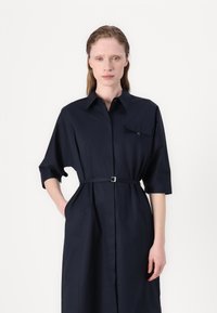 Navy blue shirt dress with a collared neckline, short sleeves, front pocket, and belted waist. Smooth fabric with a structured design.