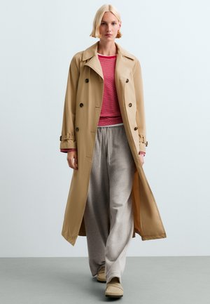 Personne blonde portant un trench-coat beige sur un haut rayé rouge et un pantalon de survêtement gris ample, marchant sur un sol gris uni avec un fond clair.