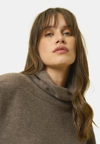 Brauner Pullover mit hohem, gefaltetem Kragen. Weiche Textur und lockere Passform. Langes, welliges Haar umrahmt das Gesicht, das mit minimalem Make-up geschmückt ist.