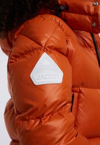 Pomarańczowa kurtka puchowa z gładkiego, błyszczącego materiału, z białą trójkątną naszywką z logo na ramieniu i pikowanym przeszyciem.