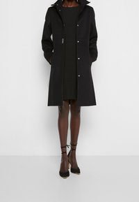 Cappotto lungo nero con colletto, design strutturato, chiusura a bottone e tasche laterali. Indossato sopra un vestito nero, completato da scarpe con tacco alto nere.