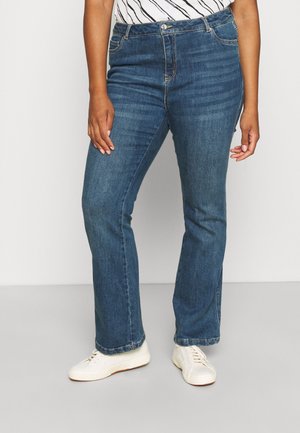 Osoba ubrana w niebieskie jeansy o prostych nogawkach, białe trampki i włożoną w spodnie pasiastą bluzkę, stoi na jasnym, jednolitym tle.