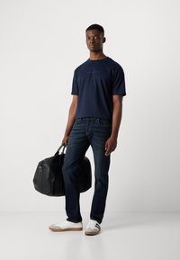 Tricou bleumarin cu mânecă scurtă, blugi închiși la culoare, adidași albi cu accente gri și o geantă mare de duffle neagră cu cusături albe.