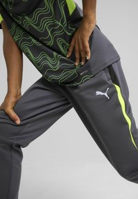 Sportska odjeća koja se sastoji od crnog gornjeg dijela s zelenim valovitim uzorcima i sivih hlača s žutim detaljem i Puma logom sa strane.