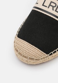Gros plan sur la pointe d’une espadrille tissée noire et beige avec des coutures visibles et un texte partiellement imprimé sur la partie supérieure en tissu.