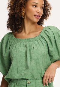 Blusa verde, con texture, maniche a sbuffo e scollatura quadrata, decorata con motivi a foglia ricamati e colletto arricciato.