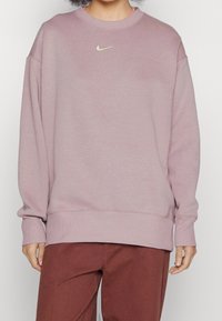 Světle růžová mikina s kulatým výstřihem, pružnými manžetami a lemem. Obsahuje malé stříbrné logo Nike na hrudi. Materiál vypadá měkký a pohodlný.