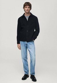 Giacca nera con colletto, in tessuto leggero, con una tasca sul petto. Abbinata a jeans azzurri chiari e scarpe nere.