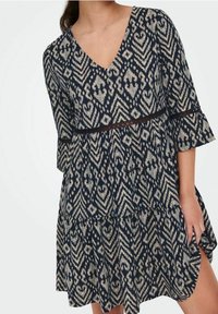 Žena v modro-béžových vzorovaných šatech s výstřihem do V, třičtvrtečními rukávy a krajkovým lemem v pase a na rukávech.