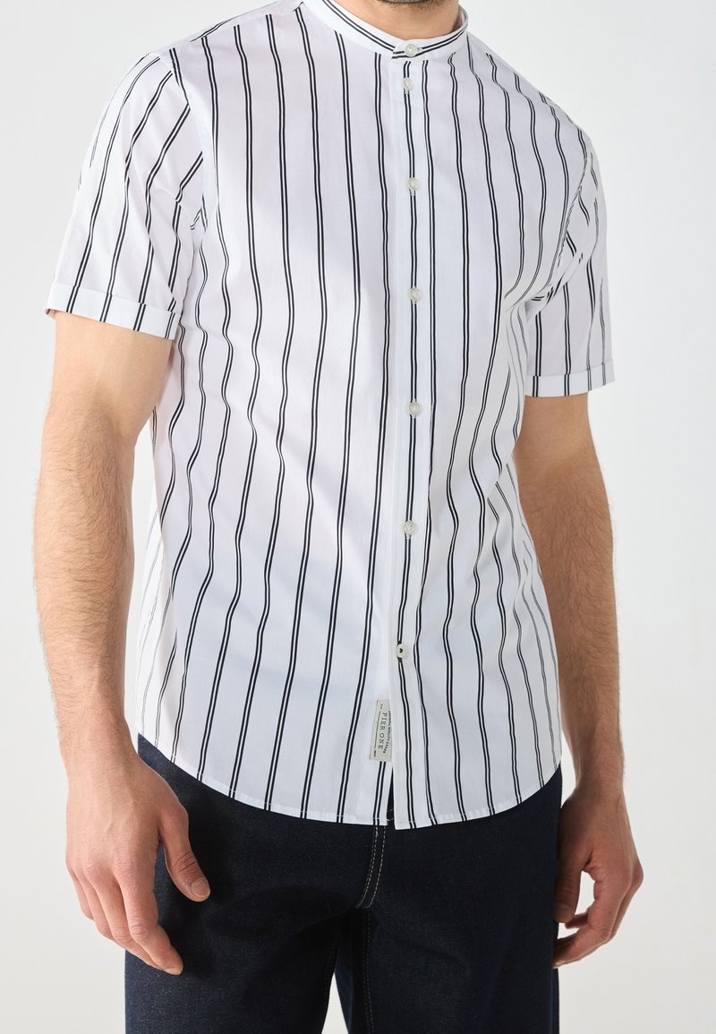 Homme portant une chemise blanche à manches courtes avec des rayures verticales noires, associée à un pantalon foncé, debout devant un fond uni.