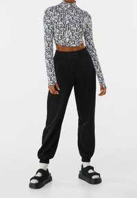 Černý zkrácený top s dlouhým rukávem a bílým písmovým vzorem, spárovaný s černými zúženými tepláky a platformovými sandály se středními pásky.