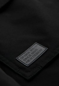 Primer plano de tela negra con una etiqueta rectangular texturizada con letras en relieve: "OMBRE WEAR DESDE 1985 NUEVO DISEÑO." Detalles de costura visibles.