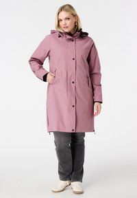 Vrouw zegt een mauve regenjas met capuchon en zwarte drukknoopjes, grijze spijkerbroek en witte sneakers terwijl ze tegen een witte achtergrond staat.