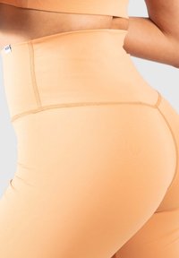 Högmidjade leggings i persikofärgad tyg, med slät yta, töjbart material och kontrasterande sömmar längs kanterna.