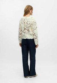 Creme hæklet top med blomster mønstre og bølget kant, parret med navy vidde bukser. Har lange ærmer og åben ryg detalje.