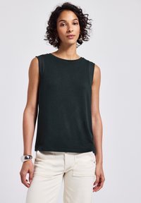 Mörkgrön ärmlös topp med rund hals och slät textur; har kontrasterande axeldetaljer. Bärs med ljusa byxor.