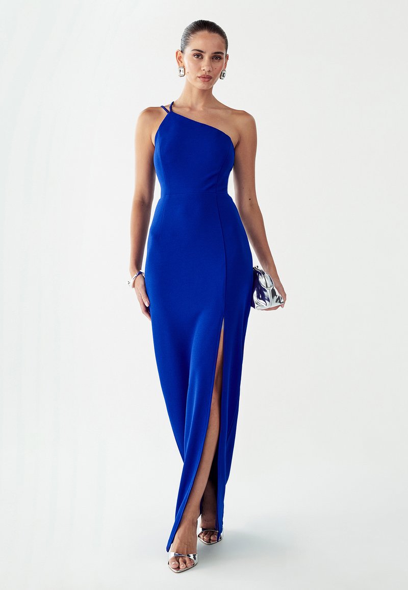 Asimetrična modra obleka s tesno prilegajočim zgornjim delom, visokim razporkom do stegna in gladko teksturo. Kombinirana s srebrno torbico in metalik petami.