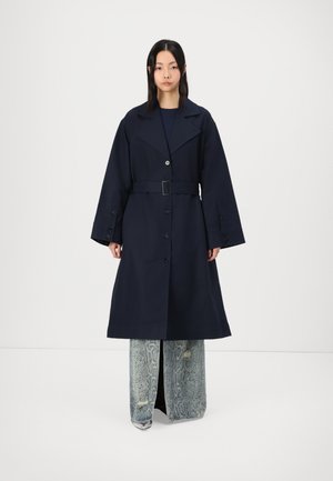 Mørkeblå trenchcoat med belte, stor krage og knapper foran, parret med vide slangeprintbukser.