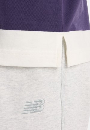 Κοντινό πλάνο του τελειώματος μοβ και λευκού πουκάμισου πάνω από ανοιχτό γκρι φόρμα με κεντημένο λογότυπο New Balance στο ύφασμα.