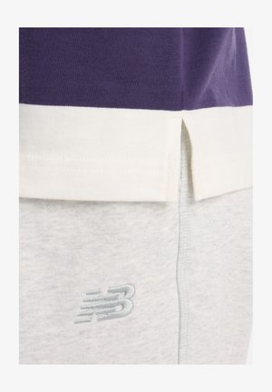 Gros plan sur l'ourlet d'une chemise violette et blanche au-dessus d'un pantalon de survêtement gris clair avec le logo New Balance brodé sur le tissu.