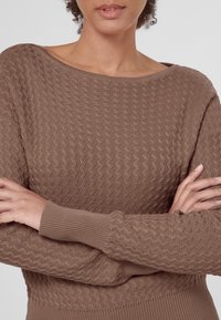 Brązowy sweter dzianinowy z teksturowanym, falistym wzorem. Posiada szeroki dekolt i ściągacze na rękawach, zaprezentowany z założonymi na sobie ramionami.